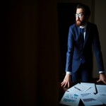 Portrait of elegant businessman in suit and eyeglasses standing by table with papers on it