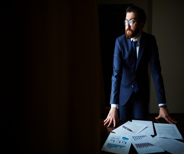 Portrait of elegant businessman in suit and eyeglasses standing by table with papers on it